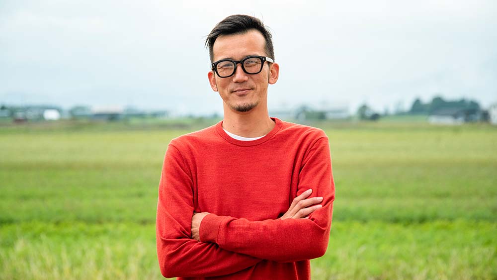 A person wearing glasses stands confidently and with crossed arms in a vast field. 